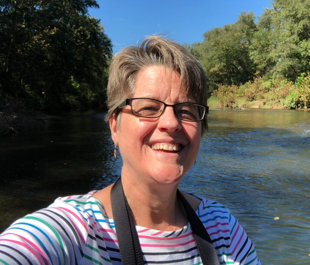 White woman with light hair and glasses in front of woods and a river.